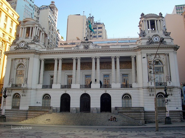 Descrição da foto para deficientes visuais: tomada da frente da câmara dos vereadores/RJ, na cor branca, mostrando a escadaria – em tom acinzentado - de acesso a porta principal