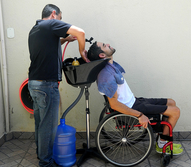 O empresário José Valente lava os cabelos do cliente no lavatório adaptável à altura da cadeira; o equipamento é levado até o local de van, junto com o galão de água, de onde sai a mangueira. Foto: Jéssica Nascimento/UOL